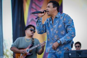 Male singer wearing a blue floral denim shirt sings into a handheld microphone on stage, with a guitarist playing a brown electric guitar nearby.