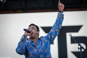 Male singer on stage wearing a blue floral shirt, microphone in hand, raising his right arm with a pointed finger during a live performance outdoors.