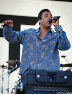Male singer wearing a blue floral shirt sings into a microphone on stage, with drums in the background, in a live performance.