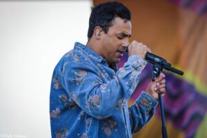 Male singer wearing a blue floral denim shirt, holding a microphone and performing on stage with a colorful background