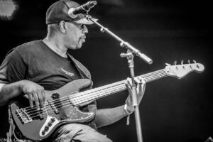 Male bassist wearing a cap, playing a bass guitar on stage with a microphone nearby.