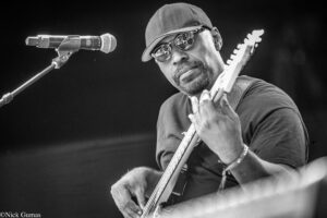 Male guitarist wearing a cap and sunglasses performs on stage, playing an electric guitar into a microphone.