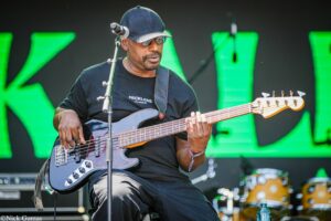 Male bass guitarist wearing a dark cap and sunglasses, playing a black bass guitar on a stage with a bright green backdrop.