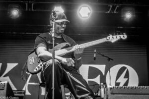 Black-and-white photo of a bass guitarist on stage, wearing a cap and sunglasses, playing a bass with a microphone in front and stage lights behind him.