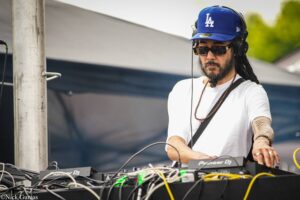 DJ wearing a blue LA cap and sunglasses performs at a mixing console outdoors, surrounded by cables and gear