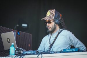 DJ wearing leopard-print cap and sunglasses, mixing tracks with headphones on a stage setup.