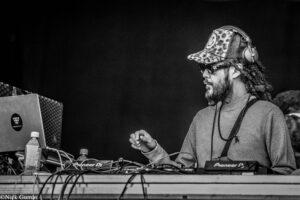 DJ wearing a leopard-print cap and sunglasses, working at a DJ mixer with cables and gear visible in front of him in a black‑and‑white shot