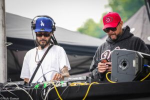 Two DJs perform at an outdoor booth with mixing equipment and many cables; left DJ wears a blue LA cap and headphones, right DJ wears a red cap and sunglasses.