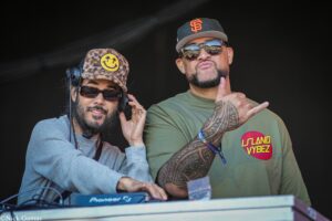 Two male DJs posing at a booth, one wearing a leopard-print hat with a yellow smiley patch and headphones, the other in a green shirt with a tattooed arm and sunglasses, giving a thumbs-up.