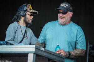 Two men wearing sunglasses and hats stand at a table with DJ gear, sharing a smile while talking to each other.