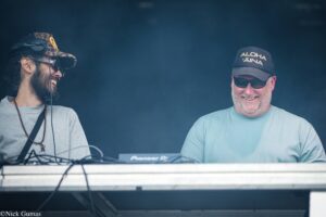 Two men DJing at a booth, smiling; left man in camouflage cap and headphones, right man wearing sunglasses and a cap that says 'ALOHA AINA'