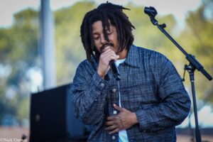 Male singer with dreadlocks performs into a handheld microphone on an outdoor stage, wearing a blue plaid shirt.