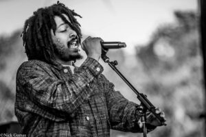 Black-and-white photo of a man with dreadlocks singing into a microphone on stage, wearing a plaid jacket.