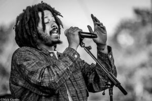 Black-and-white photo of a man with dreadlocks singing into a microphone, wearing a plaid shirt, eyes closed in performance.