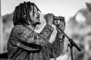 Male singer with dreadlocks, wearing a plaid shirt, singing into a handheld microphone with eyes closed on stage (black-and-white photo).