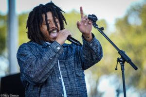 Male singer with dreadlocks performing on an outdoor stage, holding a microphone and raising his hand as he sings.