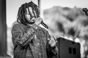 Male singer with dreadlocks singing into a handheld microphone on stage, wearing a plaid shirt and closed eyes in the moment of performance.