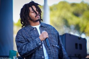 Male performer with dreadlocks holding a microphone outdoors, wearing a blue plaid denim jacket and white shirt.