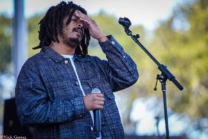 Male singer with dreadlocks on an outdoor stage, holding a microphone and resting his hand on his forehead while performing in a denim jacket.
