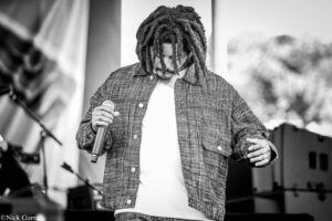 Male performer with dreadlocks holding a microphone on stage, wearing a denim jacket and white shirt.