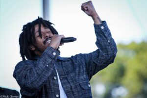 Male singer with dreadlocks raising a clenched fist while singing into a microphone outdoors, wearing a blue plaid jacket and white shirt