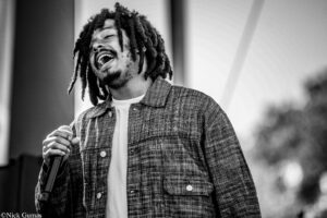 Singer with dreadlocks singing into a microphone, wearing a plaid jacket on stage (black and white)