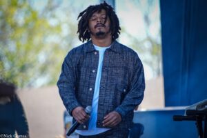 Musician with dreadlocks and denim jacket singing into a microphone on an outdoor stage, eyes closed.