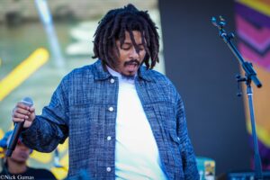 Singer with dreadlocks on stage, wearing a blue denim shirt and white tee, holding a microphone.