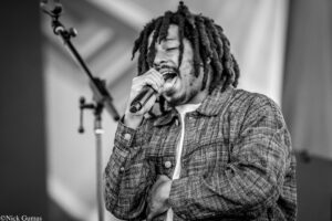 Black-and-white photo of a male singer with dreadlocks, passionately singing into a handheld microphone on stage, wearing a plaid jacket.
