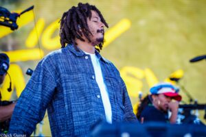 Man with dreadlocks wearing a blue denim shirt performs on stage with eyes closed.