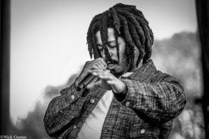 Black-and-white image of a male singer with dreadlocks singing into a microphone, wearing a plaid jacket.