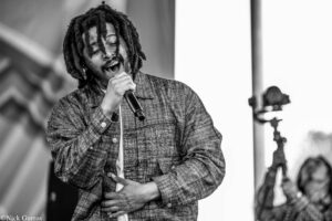 Singer with dreadlocks performs into a microphone, eyes closed, wearing a plaid jacket on stage in black and white.