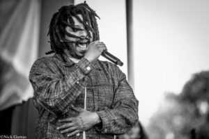Black-and-white photo of a male singer with dreadlocks passionately singing into a handheld microphone on stage, wearing a plaid shirt.