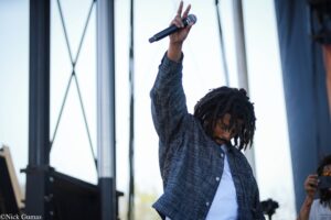 Person with dreadlocks raises a microphone on stage during a performance, wearing a denim shirt over a white tee.