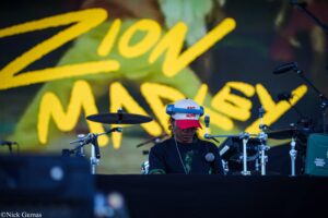 DJ wearing a white cap with red brim performing on stage, with bright yellow lettering backdrop and drums in the foreground.Material shows a concert setup with microphones and gear behind him.