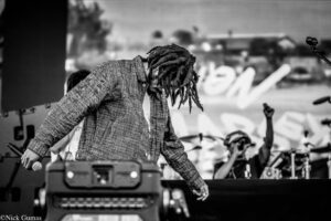 Black-and-white photo of a performer with dreadlocks leaning over a keyboard/mixer on stage, holding a microphone, with a cheering crowd in the blurred background.