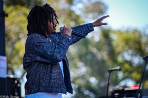 Male singer with dreadlocks performing into a handheld microphone on an outdoor stage, arm raised as he sings.