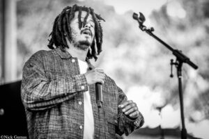 Black-and-white photo of a male singer with dreadlocks performing into a microphone on stage.