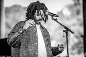 Male vocalist with dreadlocks performing on stage into a microphone, wearing a plaid jacket.