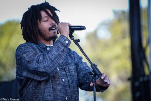Male singer with dreadlocks performing on stage, singing into a handheld mic while gripping a mic stand.