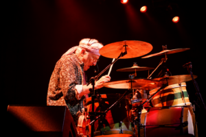 Older drummer in a patterned shirt plays a drum kit on stage under red lights, wearing a headset microphone and concentrating on the cymbals.
