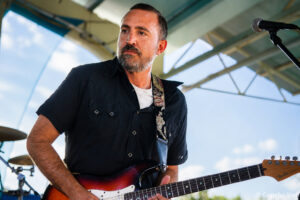 Male guitarist performing on an outdoor stage, playing a sunburst electric guitar with a patterned strap.