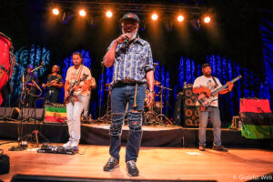 Male lead singer in plaid shirt and cap performs on a brightly lit stage with backup musicians and instruments in the background.