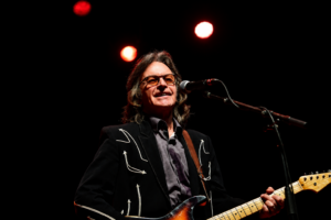 Male guitarist on stage wearing a black jacket with white embroidery, playing a sunburst electric guitar under red stage lights while singing into a mic.