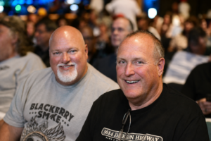 Two smiling men sit side by side at a crowded event, with a blurred audience in the background; the man on the left wears a gray Blackberry Smoke T‑shirt, the man on the right wears a black shirt and glasses hanging from his collar.