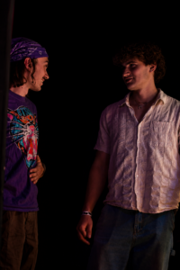 Two young men converse in a dark setting; left is in a purple bandana and graphic T-shirt, right in a light patterned shirt.