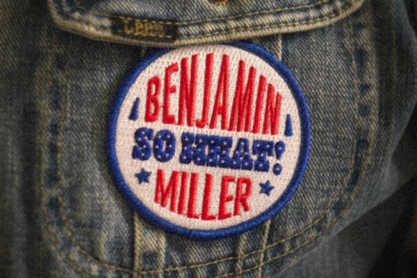 Round embroidered patch on a denim jacket reading 'BENJAMIN MILLER' with 'SO BRAVE!' in blue and red letters.