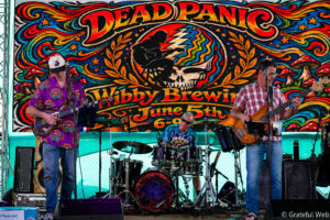 Band performing on a colorful psychedelic backdrop featuring a skull design and the word 'DEADPAN'; left guitarist in a purple shirt, center drummer, right bassist.