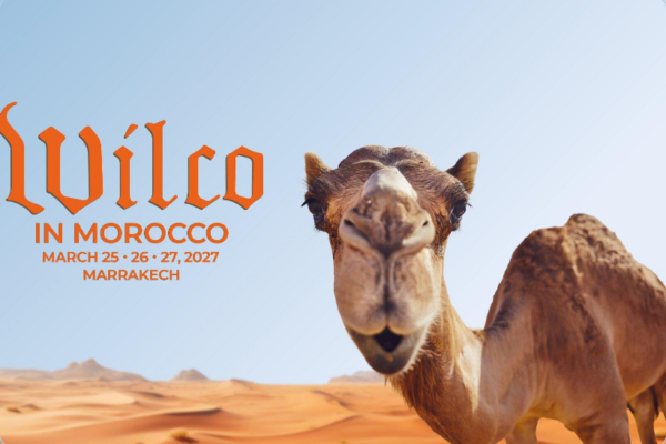 Camel in a desert landscape with dunes and a clear blue sky; event banner reads 'Wilco in Morocco' with dates March 25–27, 2027, Marrakech.