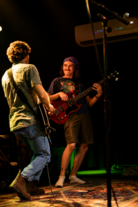 Two musicians on a dimly lit stage: a guitarist facing away in a light shirt, and a bassist with a purple bandana smiling while playing a red bass guitar.